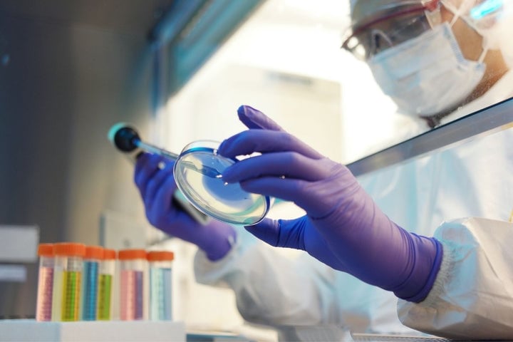 A life science lab technician working with a sample while wearing protective gloves, a face mask, goggles, and a bouffant cap.
