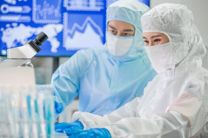 Female and male cleanroom operators working in a controlled environment, wearing hoods, face covers, full coveralls, and gloves.